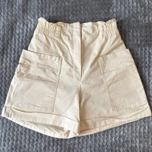 Beige högmidjade shorts från H&M - Säljer ett par beige högmidjade shorts från H&M med bred resår i midjan och stora praktiska fickor på sidorna. Shortsen har en avslappnad passform och är perfekta för varma dagar. Tillverkade i mjuk bomull för extra komfort.