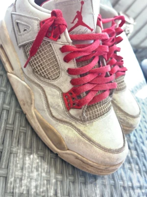 Air Jordan 4 med röda skosnören - Säljer ett par Air Jordan 4 sneakers i vitt med röda skosnören och detaljer. Skorna har meshpaneler på sidorna och tungans klassiska Jumpman-logga. Snygg retrostil och ikonisk siluett som sticker ut.