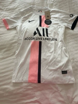 PSG vit matchtröja med rosa detalj - Snygg vit PSG matchtröja från Nike med rosa och svart rand längs framsidan. Tröjan har klubbmärke och sponsortryck på bröstet samt svarta detaljer på ärmarna. Tillverkad i lätt och ventilerande material, perfekt för fotboll eller träning.
