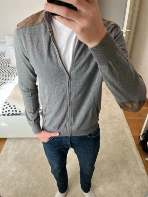 Grå ziptröja med bruna detaljer från Massimo Dutti - Grå zip tröja från Massimo Dutti med snygga läderdetaljer. Tröjan är mjukt stickad i fint skick (har några små fläckar på lädret, inget som man tänker på vid användning) —————————————————                                                             📐| Storlek: L mer som en M                                                           👖| Märke: Massimo Dutti                                      🔎 | Skick: BRA                                                         ✍🏻 | Skriv om du har några frågor