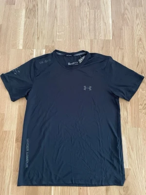 Svart Under Armour t-shirt - Snygg svart t-shirt från Under Armour med diskret logga på bröstet och tryck på ärmen. Tillverkad i ett lätt och funktionellt syntetmaterial som andas, perfekt för aktiva dagar. Klassisk passform och rund halsringning.
