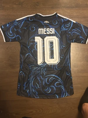 Argentina Messi matchtröja blå Adidas - Snygg Argentina matchtröja från Adidas med Messi och nummer 10 på ryggen. Tröjan är svart med blått mönster, vita detaljer och officiella AFA- och FIFA-emblem på bröstet. Korta ärmar och klassiska Adidas-ränder på axlarna. Perfekt för fotbollsfans!