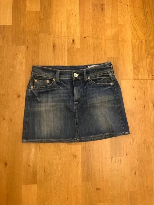Kort jeanskjol från Nolita - Säljer en snygg kort jeanskjol från Nolita med klassisk femficksdesign och slitna detaljer. Baksidan har två fickor dekorerade med färgglada nitar i zigzag-mönster. Perfekt för dig som gillar en cool och avslappnad stil.