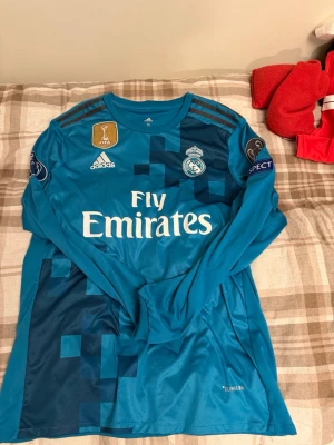Real Madrid Ronaldo matchtröja blå - Säljer en långärmad blå Real Madrid matchtröja från Adidas med Ronaldo 7 på ryggen. Tröjan har Champions League och FIFA-märken, samt Fly Emirates-tryck på bröstet. Tillverkad i funktionsmaterial och har pixelmönster i olika blå nyanser.