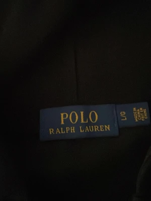 Svart hoodie från Polo Ralph Lauren - Säljer en svart hoodie från Polo Ralph Lauren med dragkedja och klassisk vit logga på bröstet. Hoodien har huva och långa ärmar, perfekt för en avslappnad och stilren look. Materialet känns mjukt och bekvämt.