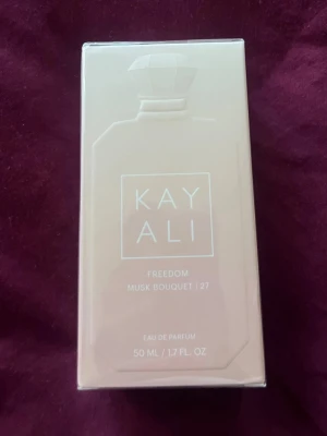 Kayali Freedom Musk Bouquet 27 EdP - Kayali Freedom Musk Bouquet 27 Eau de Parfum i 50 ml flaska. Parfymen kommer i en elegant, ljusbeige förpackning med stilren design och tydlig logotyp på framsidan. Flaskan har en klassisk form och är tillverkad av glas.