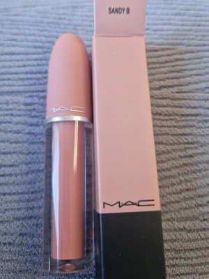 MAC Sandy B nude läppstift - MAC Sandy B är ett nude läppstift i en snygg beige-rosa ton. Hylsan är stilren med matt finish och silverdetalj, och själva läppstiftet har en klassisk rundad form. Perfekt för dig som gillar naturliga färger och vill ha ett trendigt tillskott i sminkväskan.