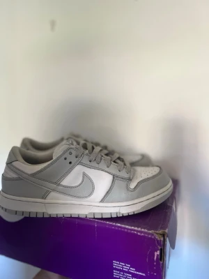 Nike Dunk Low grå/vit sneakers - Nike Dunk Low sneakers i grått och vitt med klassisk swoosh på sidan. Skorna har lågt skaft, perforerad tåbox och snörning. Tillverkade i läder med en vit sula och grå detaljer. Perfekta för dig som gillar stilrena och tidlösa sneakers.