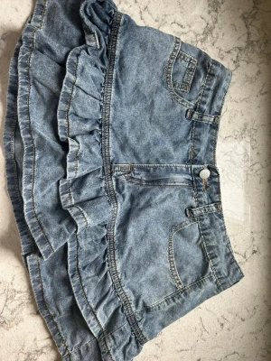 Jeansskort med volanger i denim - Supersöt jeansskort i ljusblå denim med volangdetaljer runt om. Klassisk femficksmodell med dragkedja och knapp framtill. Perfekt för dig som vill ha en lekfull och trendig look. Materialet är 100% bomullsdenim.