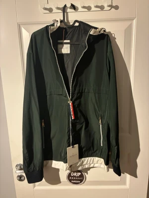 Grön vindjacka med huva från Moncleer - Snygg mörkgrön vindjacka från Moncler med vit huva och vita detaljer. Jackan har dragkedja framtill, två fickor med dragkedja och svarta muddar vid ärmsluten. Tillverkad i lätt och tåligt material, perfekt för blåsiga dagar.