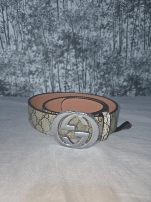 Gucci GG beige bälte med silverdetalj - Snyggt beige bälte från Gucci med klassiskt GG-mönster och stor silverfärgad GG-spänne. Bältet har en lyxig känsla och är tillverkat i skinn med detaljer i silver. Perfekt accessoar för att lyfta din outfit.