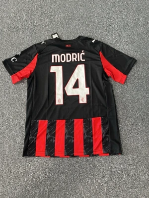 AC Milan matchtröja Modrić #14 - Snygg AC Milan matchtröja från Puma med Modrić och nummer 14 på ryggen. Tröjan är svart och röd med klassiska ränder, korta ärmar och klubbmärke på bröstet. Tillverkad i lätt och ventilerande material, perfekt för fotboll eller träning.