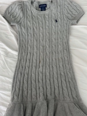 Grå stickad klänning med volang - Supersöt grå stickad klänning från Ralph Lauren med kabelstickat mönster och volangkant nedtill. Klänningen har korta ärmar och en liten broderad logga vid bröstet. Perfekt för dig som gillar klassisk och mysig stil.