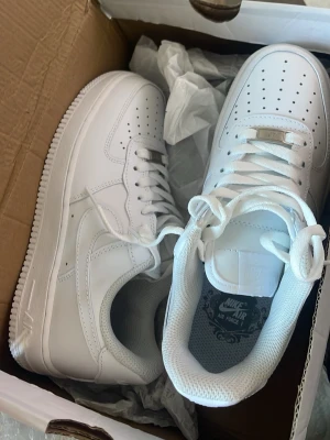 Nike Air Force 1 vita sneakers - Klassiska helvita Nike Air Force 1 sneakers i läder med perforerad tåbox, vita skosnören och diskret Nike-logga på sidan. Tidlös och clean design som passar till allt. 