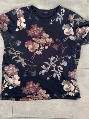 Blommig t-shirt från Selected Homme - Mörkblå t-shirt från Selected Homme med stora blommiga mönster i vinrött, beige och vitt. Klassisk rund halsringning och korta ärmar. Tillverkad i mjuk bomull för en bekväm känsla. Perfekt för dig som vill sticka ut med en snygg och trendig look.
