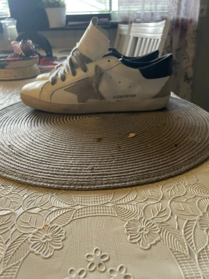 Vita Golden Goose sneakers med stjärna - Snygga vita Golden Goose sneakers med grå stjärna på sidan och svart hälparti. Skorna har grå skosnören och klassisk låg profil. Tillverkade i läder med slitna detaljer för en trendig look. Perfekta för dig som gillar streetstyle och vill sticka ut. Skorna är hyfsat tighta för dem som har breda eller tjocka fötter.
