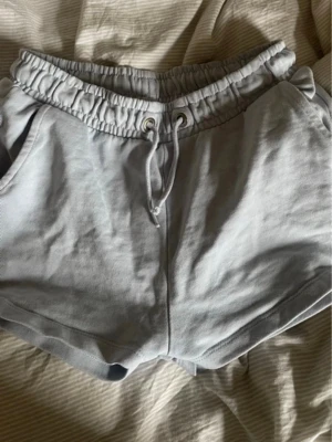 Ljusgrå mjukisshorts med snörning - Säljer ett par ljusgrå mjukisshorts med resårmidja och snörning framtill. Shortsen har två sidofickor och är gjorda i mjukt bomullsmaterial. Perfekta för chill dagar eller träning.