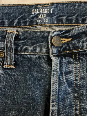 Blå raka jeans från Carhartt WIP - Säljer ett par klassiska blå jeans från Carhartt WIP i modellen 28/32. Jeansen har raka ben, snygga slitningar och kontrastsömmar. Tillverkade i slitstark denim med en cool vintagekänsla och metallknapp med Carhartt-logga.