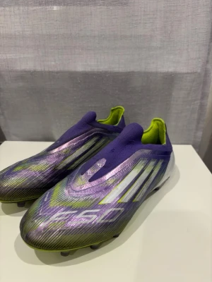 Lila Adidas X Speedflow fotbollsskor - Säljer ett par Adidas X Speedflow fotbollsskor i lila och neongrönt med vita detaljer. Skorna har en strömlinjeformad design utan snörning och ovandel i syntetmaterial för lätt vikt och bra bollkontroll. Perfekta för dig som vill ha snabbhet och stil på planen.