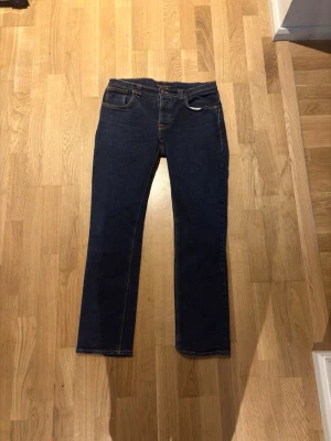 Mörkblå raka jeans från Nudie, Grim Tim 32L 36W - Säljer ett par mörkblå jeans från Nudie med klassisk rak passform. Modell grim tim 32L 36 W. Jeansen har orangea kontrastsömmar och är tillverkade i ett denimtyg. Perfekta för dig som gillar en tidlös och clean look.