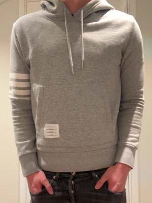 Thom browne hoodie - Snygg grå hoodie från Thom Browne med vita ränder på vänster ärm och klassisk magficka. Dragsko i huvan och ribbade muddar. Liten vit etikett framtill ger en clean och modern look. Perfekt för dig som gillar stilrena detaljer.