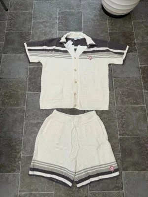 Stickat set i vitt och grått från Casablanca  - Säljer ett stickat set med kortärmad kofta och shorts i vitt och grått. Koftan har knappar, krage och två fickor framtill. Shortsen har dragsko i midjan och fickor. Båda delarna har randiga detaljer och en liten röd logotyp. Perfekt för en avslappnad look.
