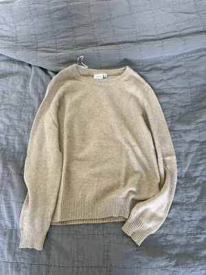 Beige stickad tröja  - En stilren beige stickad tröja  med rund halsringning och ribbade muddar vid ärmslut och nederkant. Tröjan har en klassisk passform och är perfekt för lager på lager. Materialet ser ut att vara mjukt och bekvämt.