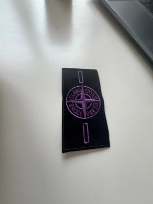Stone Island badge - purple - Säljer en svart patch från Stone Island med broderad logga i lila. Klassisk rektangulär form och detaljerad kompassdesign. Perfekt att fästa på jacka eller tröja för att ge din outfit en unik touch.