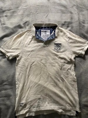Vit piké från Morris med brodyr - Säljer en vit pikétröja från Morris med broderad logga på bröstet och snygga blå blommiga detaljer i kragen. Klassisk krage och korta ärmar. Perfekt för dig som gillar stilrena plagg med lite extra detaljer.