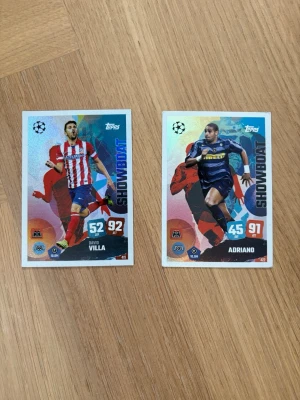 Fotbollssamlarkort Showboat Topps - Två fotbollssamlarkort från Topps med spelarna David Villa och Adriano. Korten har färgstarka motiv, holografisk yta och texten 'SHOWBOAT'. Perfekt för dig som gillar att samla på fotbollskort eller vill ha något unikt till din samling.