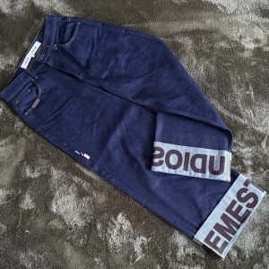 Mörkblå raw denims från EME Studios - Säljer ett par mörkblå jeans från EME Studios med breda uppvik där det står 'EMESTUDIOS' i stora bokstäver. Klassisk femficksmodell med raka ben och kontrastsömmar. Snygg detalj med liten flagga på sidan. Perfekta för en trendig streetwear-look.