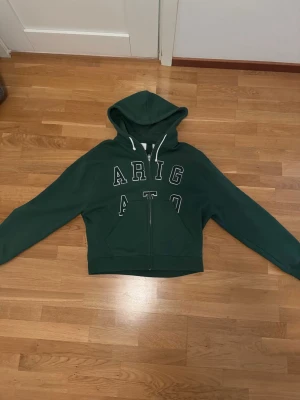 Grön zip hoodie från Axel Arigato - Mörkgrön hoodie från Axel Arigato med dragkedja framtill och vita snören i huvan. Stor logga i vitt på bröstet. Tillverkad i 100% ekologisk bomull för en skön och hållbar känsla. Perfekt för en avslappnad och trendig look. Säljer på grund av att jag inte använder den mer.