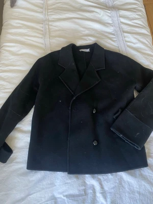 Svart ullkappa från Marville Road - Stilren svart kappa från Marville Road i ullmaterial. Klassisk dubbelknäppt design med breda slag och raka ärmar. Perfekt för dig som gillar en tidlös och enkel look med en touch av elegans.