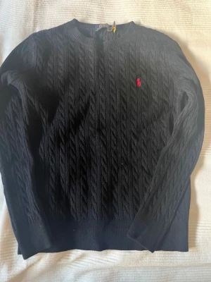 Svart kabelstickad tröja Polo Ralph Lauren - Kabelstickad svart tröja från Polo Ralph Lauren med rund halsringning och den klassiska röda logotypen broderad på bröstet. Tröjan har långa ärmar och ribbade muddar vid ärmslut och nederkant. Perfekt för en stilren och avslappnad look.