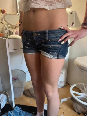 Mörkblå denimshorts med nitar - Säljer ett par mörkblå denimshorts med låg midja och råa kanter. Shortsen har dekorativa nitar och flera fickor både fram och bak. Perfekta för sommaren och ger en cool, avslappnad look. Kanterna måste vikas upp själv!!