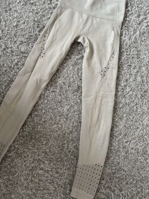 Beige seamless tights från Gymshark - Beige tights från Gymshark med seamless design och hög midja. Tightsen har snygga meshdetaljer längs benen och logga på höften. Materialet är stretchigt och andas, perfekt för gym och träning. De är knappt använda.