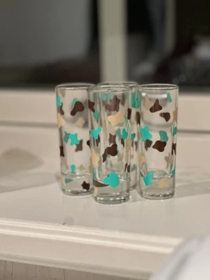 Fyra shot glas med färgglatt mönster - Säljer fyra shot glas med unikt mönster i turkos, beige och brunt. Glasen är cylinderformade och har ett lekfullt, abstrakt tryck runt hela ytan. Perfekta för drycker eller som dekoration i köket. 6cl bör hand diskas 