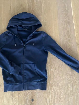 Mörkblå zip hoodie från Polo Ralph Lauren - Snygg mörkblå hoodie från Polo Ralph Lauren med dragkedja framtill och klassisk logga broderad på bröstet. Hoodien har huva med dragsko och två fickor framtill. Tillverkad i mjukt bomullsmaterial som känns skönt mot huden.