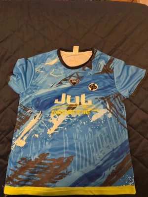JUL T-shirt  - Snygg blå sporttröja från Jul Decennie med grafiskt tryck i vitt, svart och gult. Tröjan har rund hals med svart kant, korta ärmar och tryck både fram och bak med text och loggor. Tillverkad i ett lätt och snabbtorkande material, perfekt för träning.