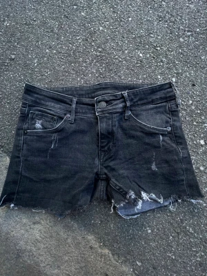 Svarta jeansshorts med rå kant - Säljer ett par svarta jeansshorts med rå kant och slitna detaljer. Klassisk femficksmodell med knapp och dragkedja framtill. Perfekta för dig som gillar en edgy och avslappnad stil.