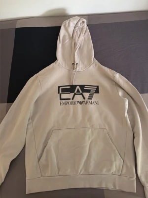 Beige hoodie från Emporio Armani - Snygg beige hoodie från Emporio Armani med stor EA7-logga på bröstet. Hoodien har huva med dragsko och en stor magficka framtill. Tillverkad i mjukt material som känns skönt mot huden. Perfekt för en avslappnad och trendig look.