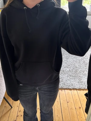 Svart hoodie med ficka framtill - En klassisk svart hoodie med huva och dragsnören. Hoodien har en stor känguruficka framtill och ribbade muddar vid ärmslut och nederkant. Perfekt för en avslappnad och stilren look. 