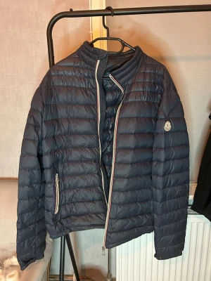 Moncler Daniel dunjacka  - Snygg mörkblå dunjacka från Moncler med quiltad design och ståkrage. Jackan har dragkedja framtill med röd, vit och blå kant, samt två fickor med dragkedja. Moncler-logga på vänster ärm. Perfekt för kalla vinterdagar. Size 4. Bra skick utom att dragkedjan börjar fucka lite