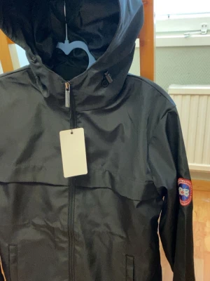 Svart vindjacka med luva och patch - Snygg svart vindjacka med luva och dragkedja framtill. Jackan har en patch på ena ärmen och två sidofickor. Tillverkad i ett lätt och vattenavvisande material, perfekt för blåsiga dagar. Enkel och stilren design som passar till det mesta.