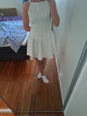 Vit kort klänning från Zara - Säljer en vit kort klänning från Zara med figurnära överdel, knappar framtill och utställd kjol. Klänningen är ärmlös och har en hög halsringning. Perfekt för sommarens alla tillfällen!❤️‍🔥