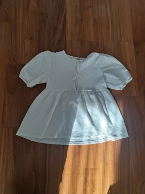 Vit blus med puffärm och rosetter i fram - Romantisk vit blus med puffiga kortärmar och två söta knyt framtill. Blusen har en flowy volang nedtill och ett lätt strukturerat tyg som ger en luftig känsla. Perfekt för dig som gillar stilrena och feminina detaljer.❤️