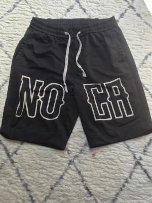 Svarta shorts med tryck och snörning - Säljer ett par svarta shorts med vita snören i midjan och stort vitt tryck med texten 'NO CA' på framsidan. Shortsen har elastisk midja och är tillverkade i mjukt material, perfekta för avslappnade dagar.
