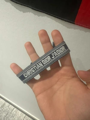 Christian Dior J'Adior armband - Snyggt armband från Christian Dior med texten 'J'Adior' i vitt på en svart och grå bakgrund. Armbandet är vävt i tyg och har en stilren, modern look som passar perfekt till din outfit.
