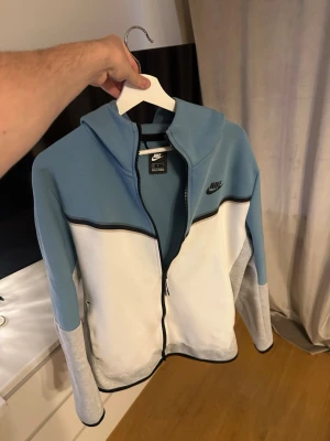 Nike tech zip  - Nike hoodie i blått och vitt med svarta detaljer och dragkedja framtill. Tröjan har huva, svarta loggan på bröstet och gråmelerade ärmar. Tillverkad i mjukt material som känns skönt mot huden. Perfekt för en avslappnad och sportig stil.