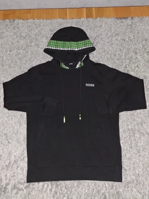 Svart hoodie från BOSS med grön vitt detalj - Snygg svart hoodie från BOSS med grön och vit logotypdetalj på huvan och dragsko med gröna ändar. Klassisk passform med ficka framtill och broderad BOSS-logga på bröstet. Perfekt för en chill och trendig look.                                                        Ups! På den sista bilden är hoodie lite sliten av tvätten men syns sällan. Om det finns frågor om den gärna skriv till mig!